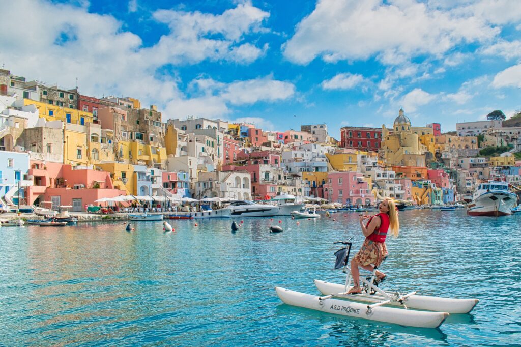 Woman on a water bike in the port of Marina Corricella di Procida with the characteristics colorful houses in the background on a waterbike