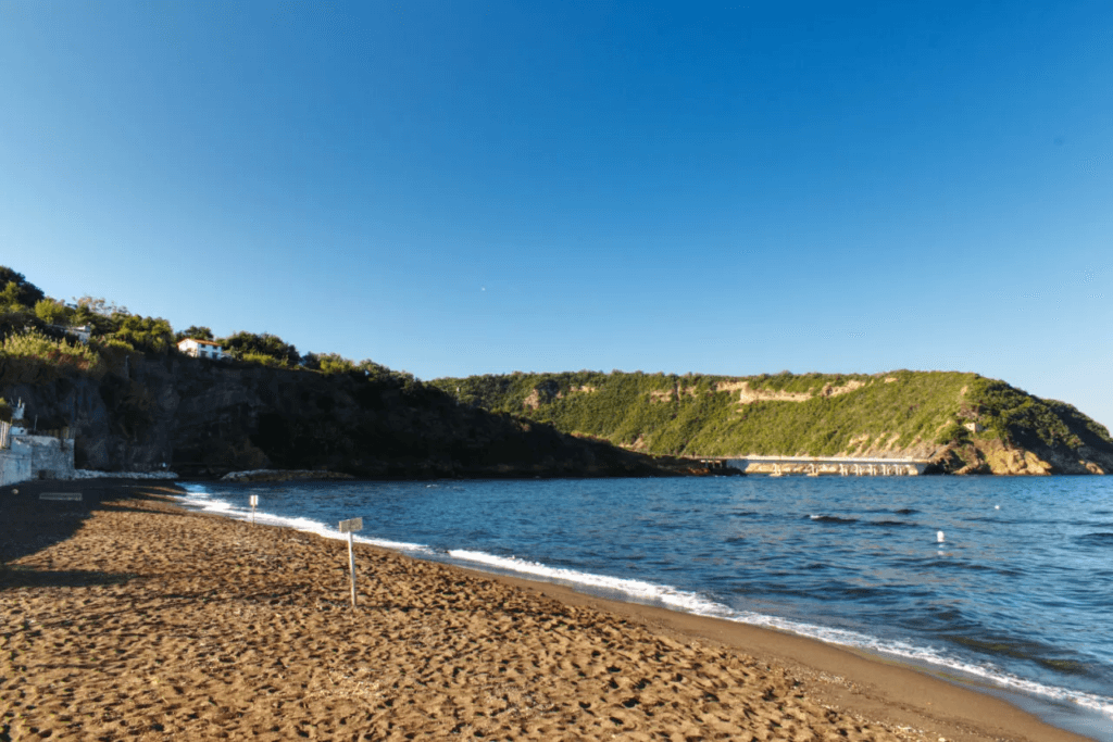 Spiaggia di sabbia scura con mare blu e colline verdi sotto un cielo limpido