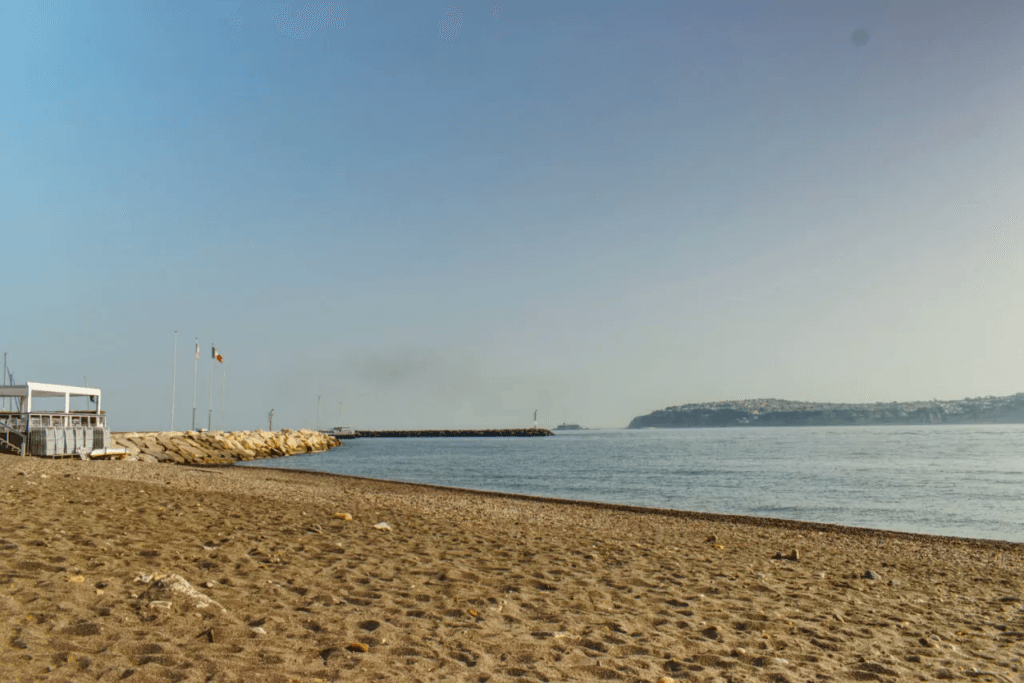 Spiaggia sabbiosa con un molo in pietra, bandiere e una città costiera in lontananza sotto un cielo sereno